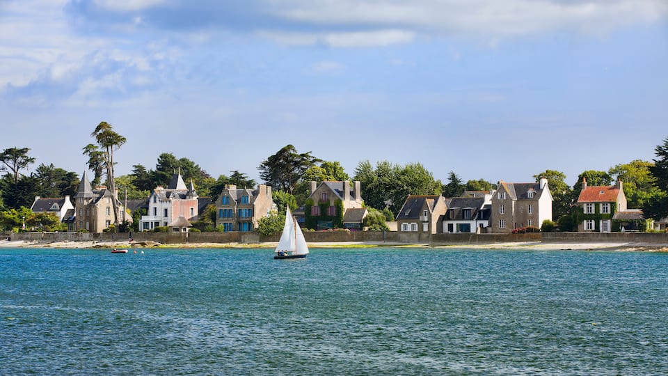 A coastal town with historic houses, a sailboat on the water, and a clear sky.