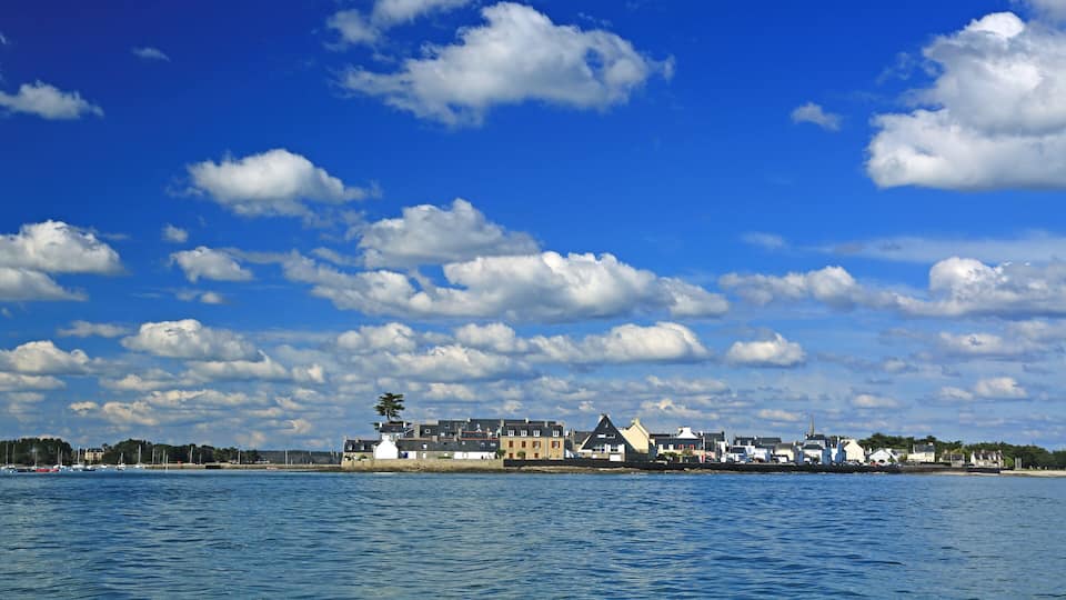 A coastal town with buildings along the shore, a clear blue sky, and scattered clouds.