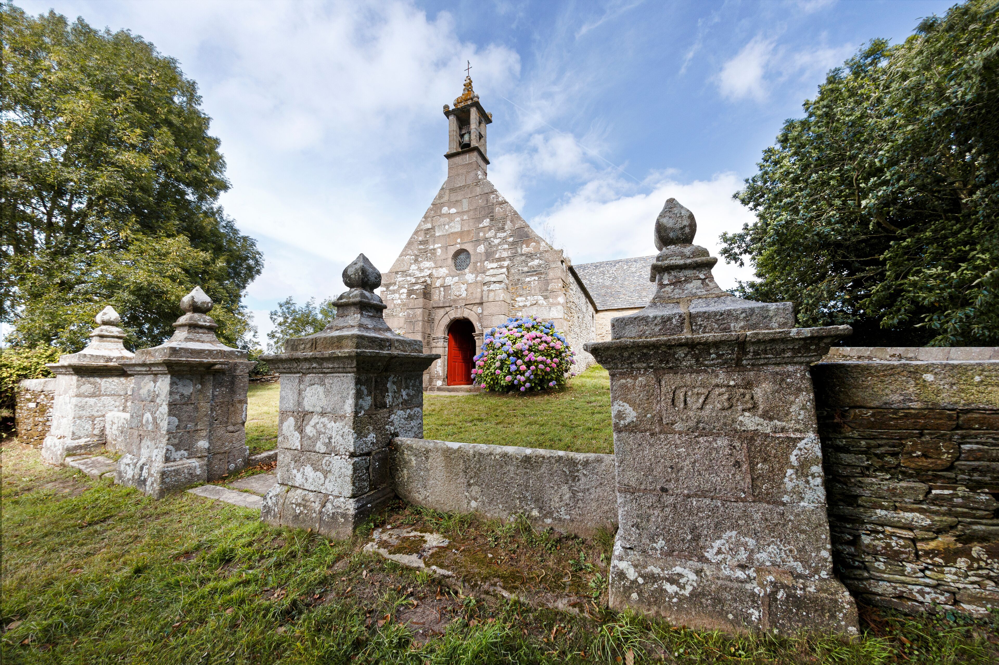 A stone church with a red door, surrounded by stone walls and a flower bed.