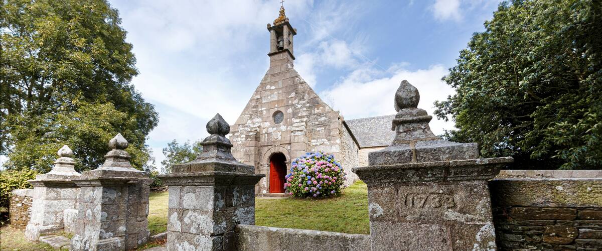 A stone church with a red door, surrounded by stone walls and a flower bed.