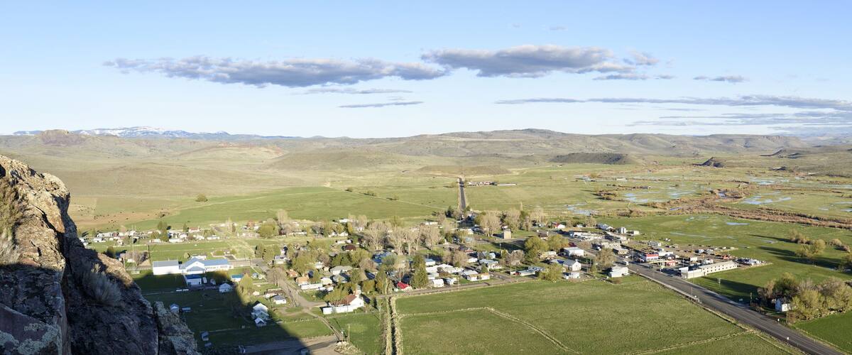 A rural town nestled in a valley with green fields, a river, and distant mountains.