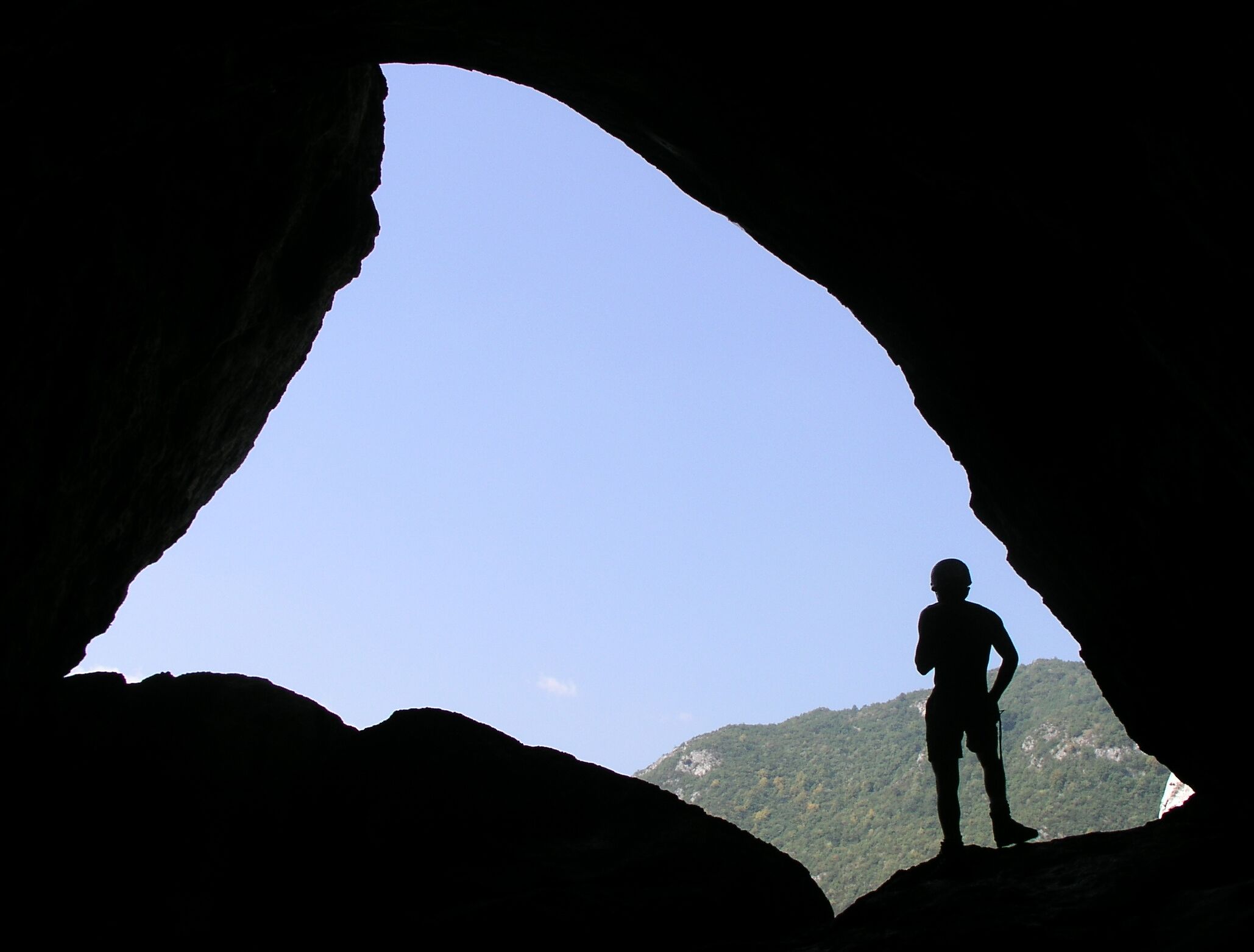 A person silhouetted against a clear sky, standing in a natural rock formation with a mountain in the background.