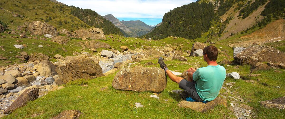 A man sitting on a rock in a mountainous area with green grass and scattered rocks.