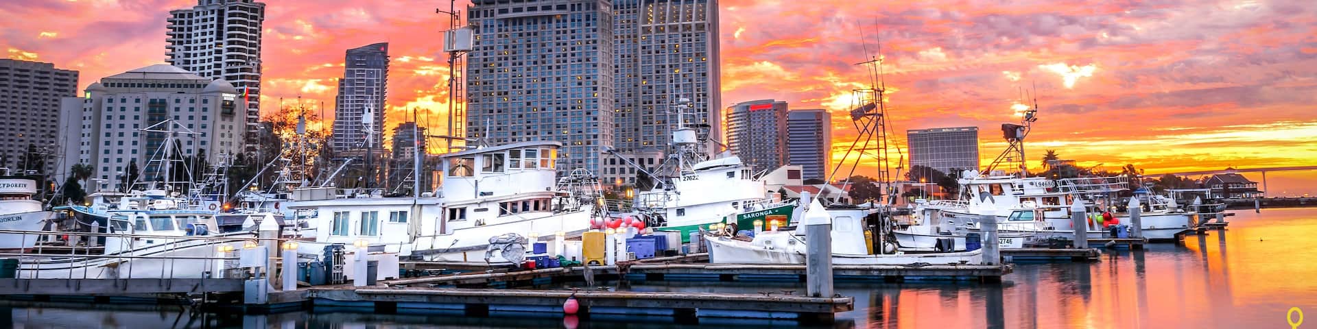 A marina with boats docked, a city skyline with high-rise buildings, and a sunset with pink and orange clouds.