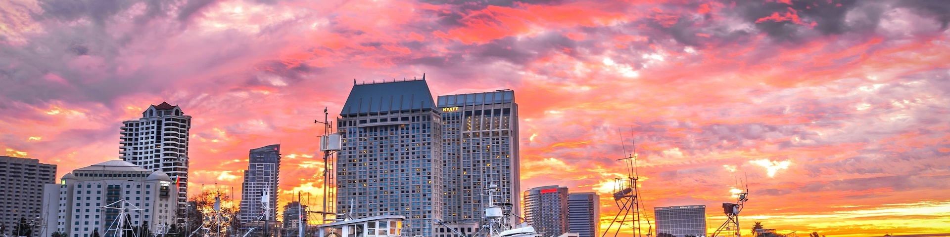 Uma marina com barcos atracados, um horizonte urbano com edifícios altos e um pôr do sol com nuvens rosadas e alaranjadas.