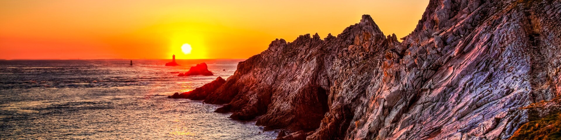 Un atardecer sobre una costa rocosa con un faro a lo lejos.
