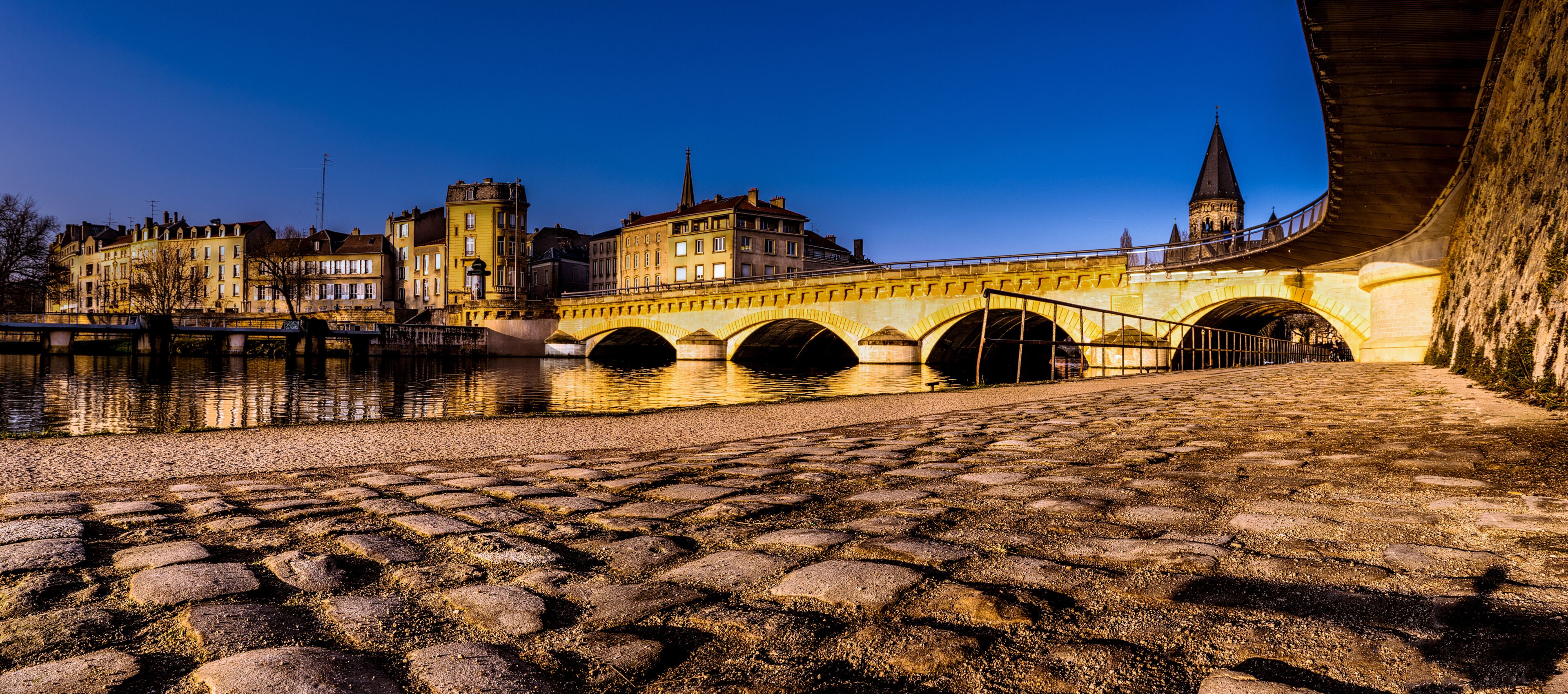 Un pont de pierre enjambe une rivière, avec des bâtiments historiques et le clocher d’une église en arrière-plan.