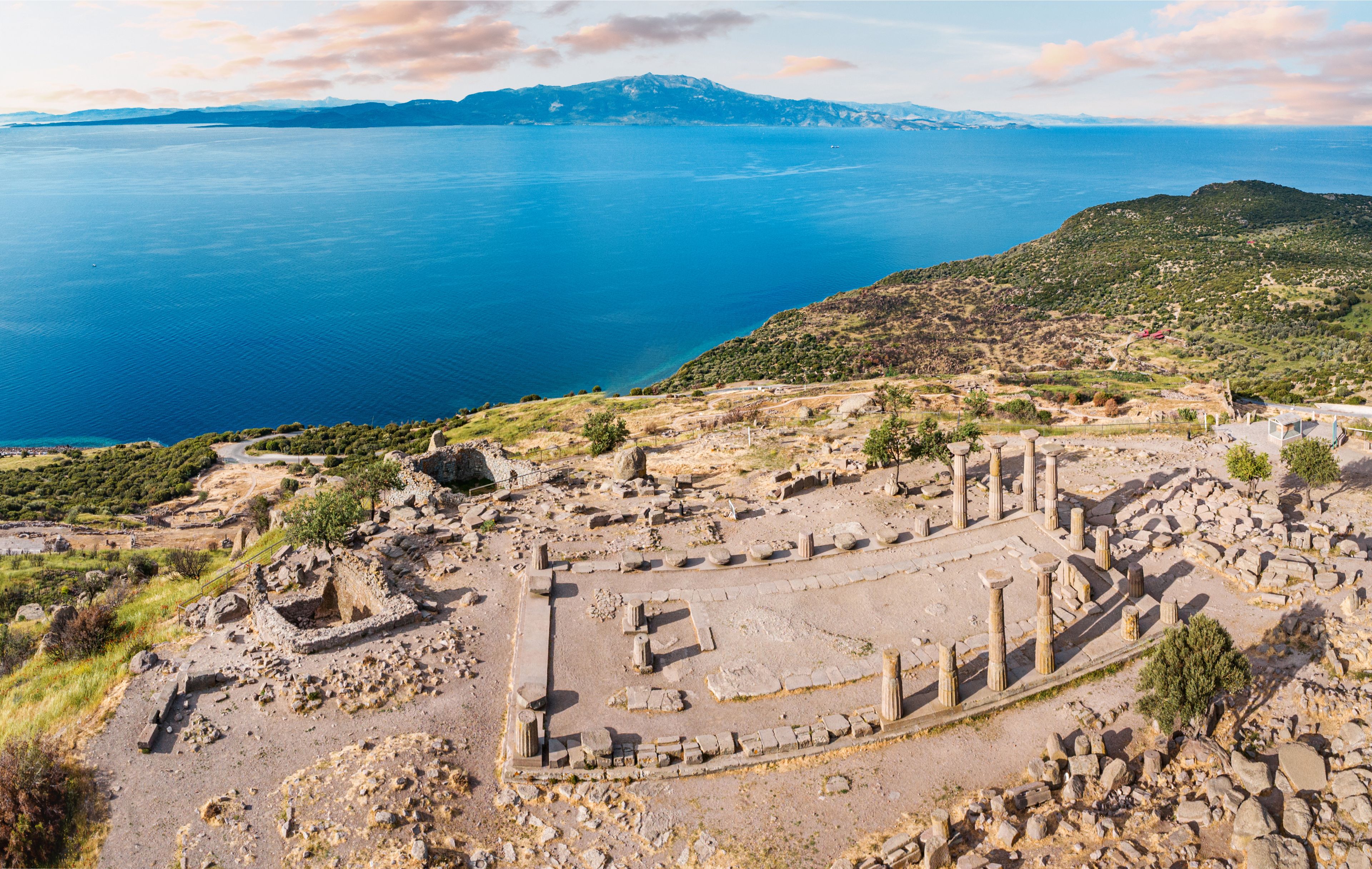 Des ruines antiques, avec des colonnes et un théâtre, dominant une mer bleue et des montagnes lointaines.