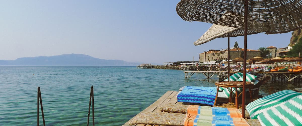 A wooden pier with lounge chairs and a thatched umbrella overlooking a calm sea with distant mountains.
