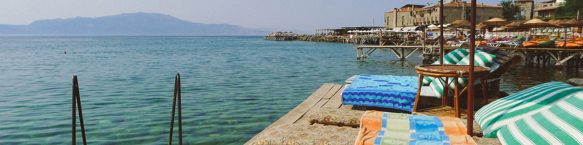 A wooden pier with lounge chairs and a thatched umbrella overlooking a calm sea with distant mountains.
