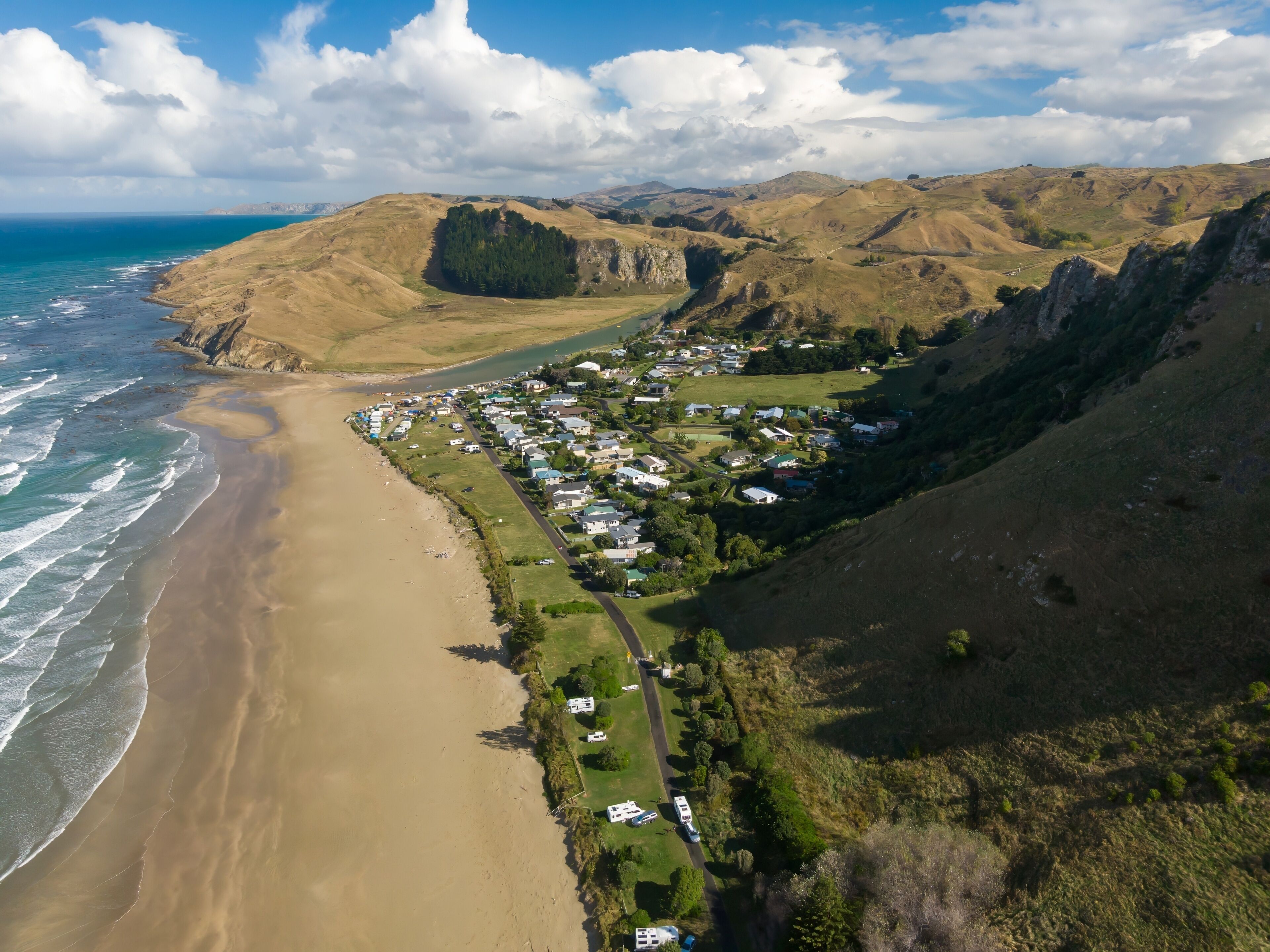 Aerial view of Kairakau Beach, Hawke's Bay, New Zealand, showing the beach, ocean, and a small coastal town nestled between rolling hills. Tourists visit for recreation.