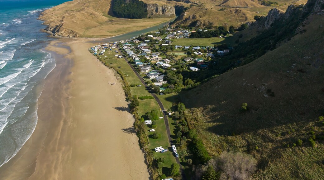 Aerial view of Kairakau Beach, Hawke's Bay, New Zealand, showing the beach, ocean, and a small coastal town nestled between rolling hills. Tourists visit for recreation.
