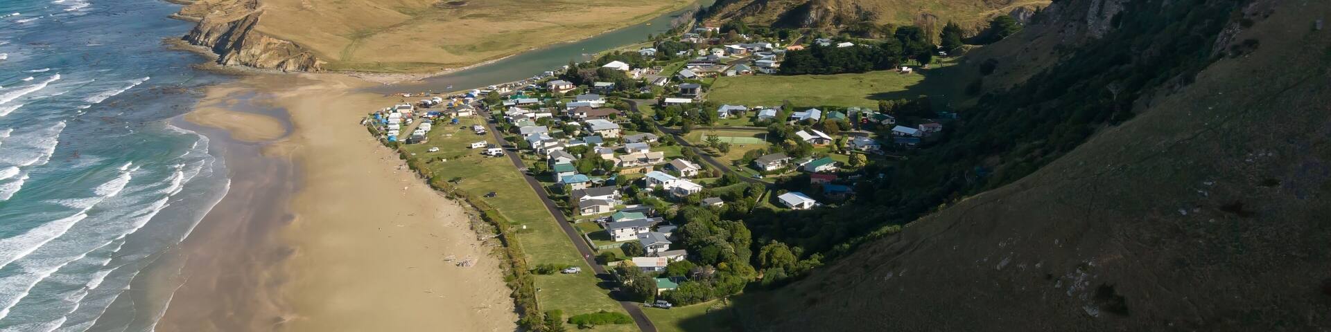 Aerial view of Kairakau Beach, Hawke's Bay, New Zealand, showing the beach, ocean, and a small coastal town nestled between rolling hills. Tourists visit for recreation.