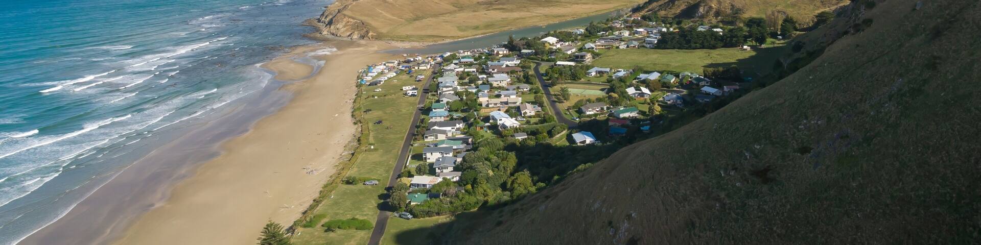 Aerial view of Kairakau Beach, Hawke's Bay, New Zealand, showing the beach, ocean, and a small coastal town nestled between rolling hills. Tourists visit for recreation.