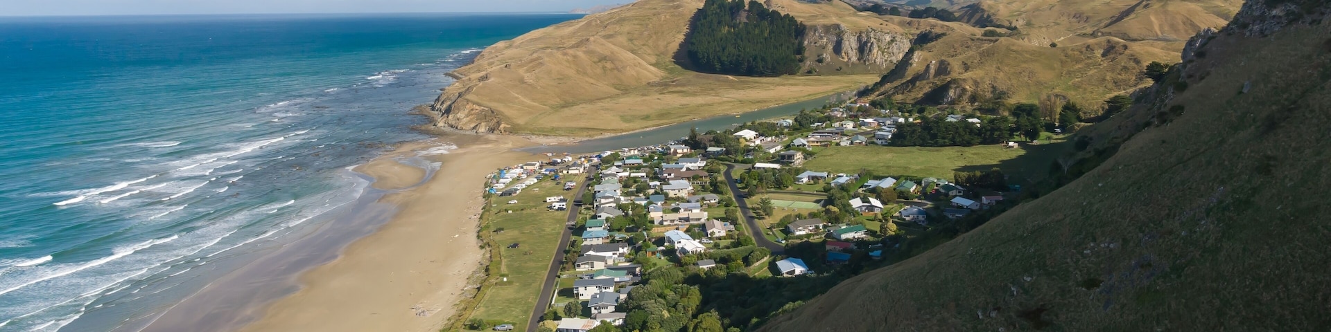 Aerial view of Kairakau Beach, Hawke's Bay, New Zealand, showing the beach, ocean, and a small coastal town nestled between rolling hills. Tourists visit for recreation.