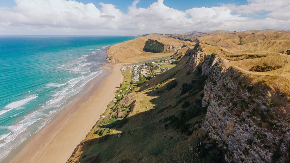 Aerial view of Kairakau Beach, Hawke's Bay, New Zealand, showing the beach, ocean, and a small coastal town nestled between rolling hills. Tourists visit for recreation.
