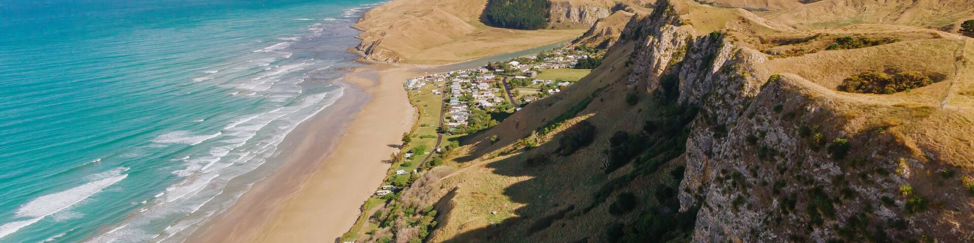 Aerial view of Kairakau Beach, Hawke's Bay, New Zealand, showing the beach, ocean, and a small coastal town nestled between rolling hills. Tourists visit for recreation.