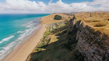 Aerial view of Kairakau Beach, Hawke's Bay, New Zealand, showing the beach, ocean, and a small coastal town nestled between rolling hills. Tourists visit for recreation.