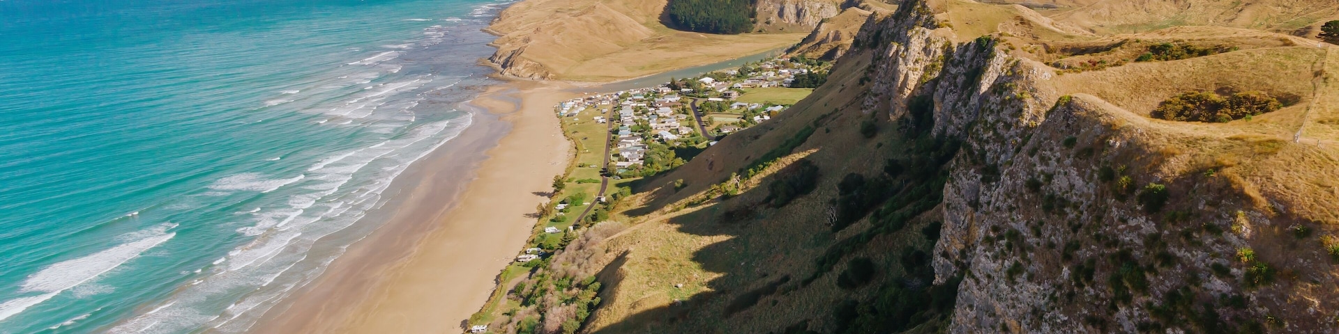 Aerial view of Kairakau Beach, Hawke's Bay, New Zealand, showing the beach, ocean, and a small coastal town nestled between rolling hills. Tourists visit for recreation.