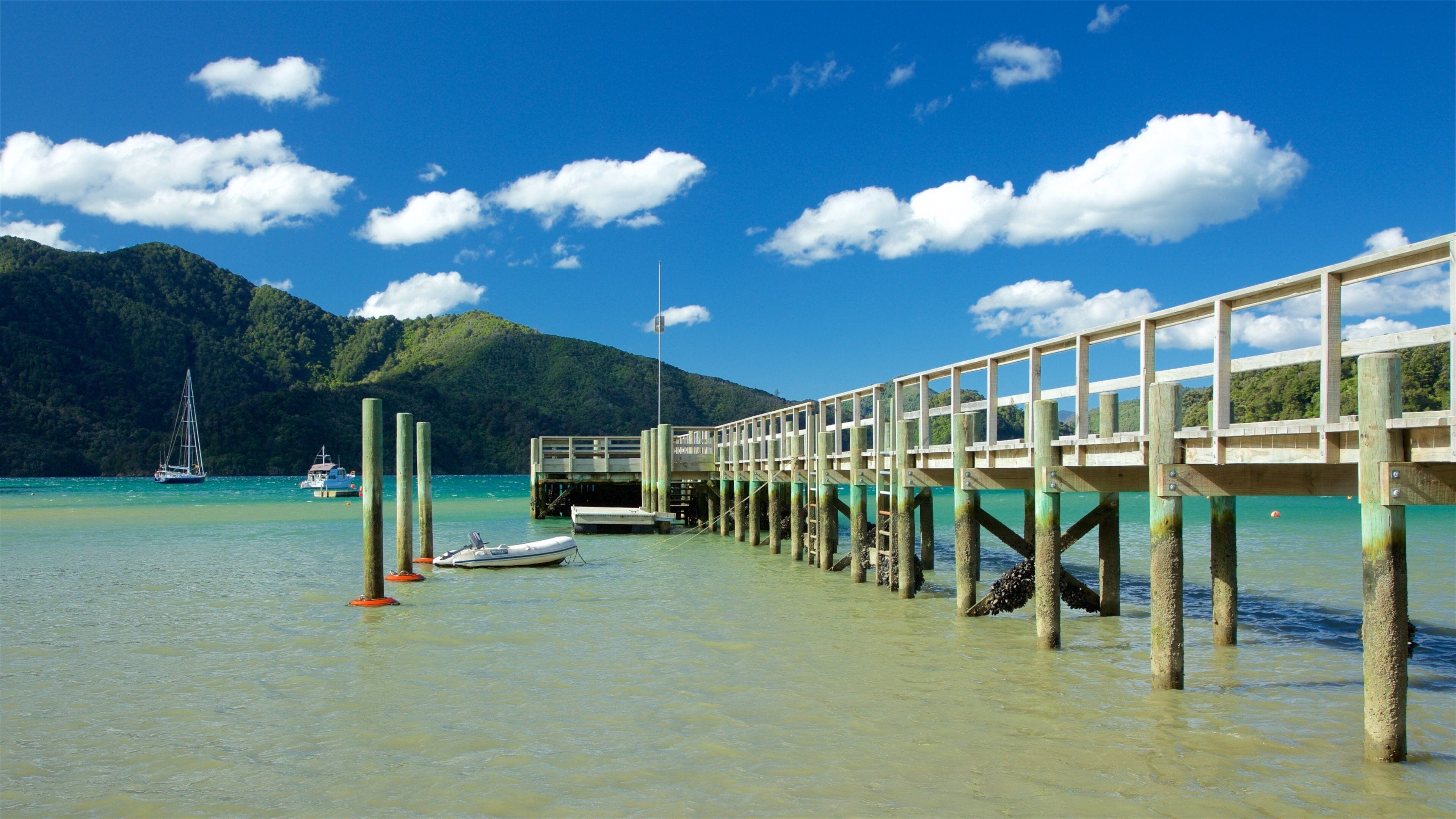 A wooden pier extending into turquoise waters with boats and a sailboat in the distance, surrounded by lush green hills.