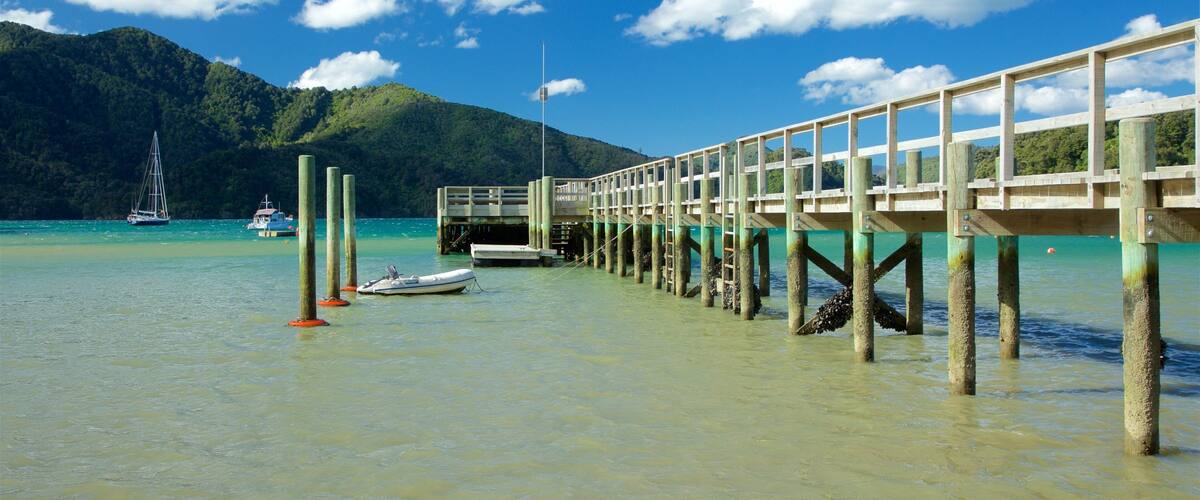 A wooden pier extending into turquoise waters with boats and a sailboat in the distance, surrounded by lush green hills.
