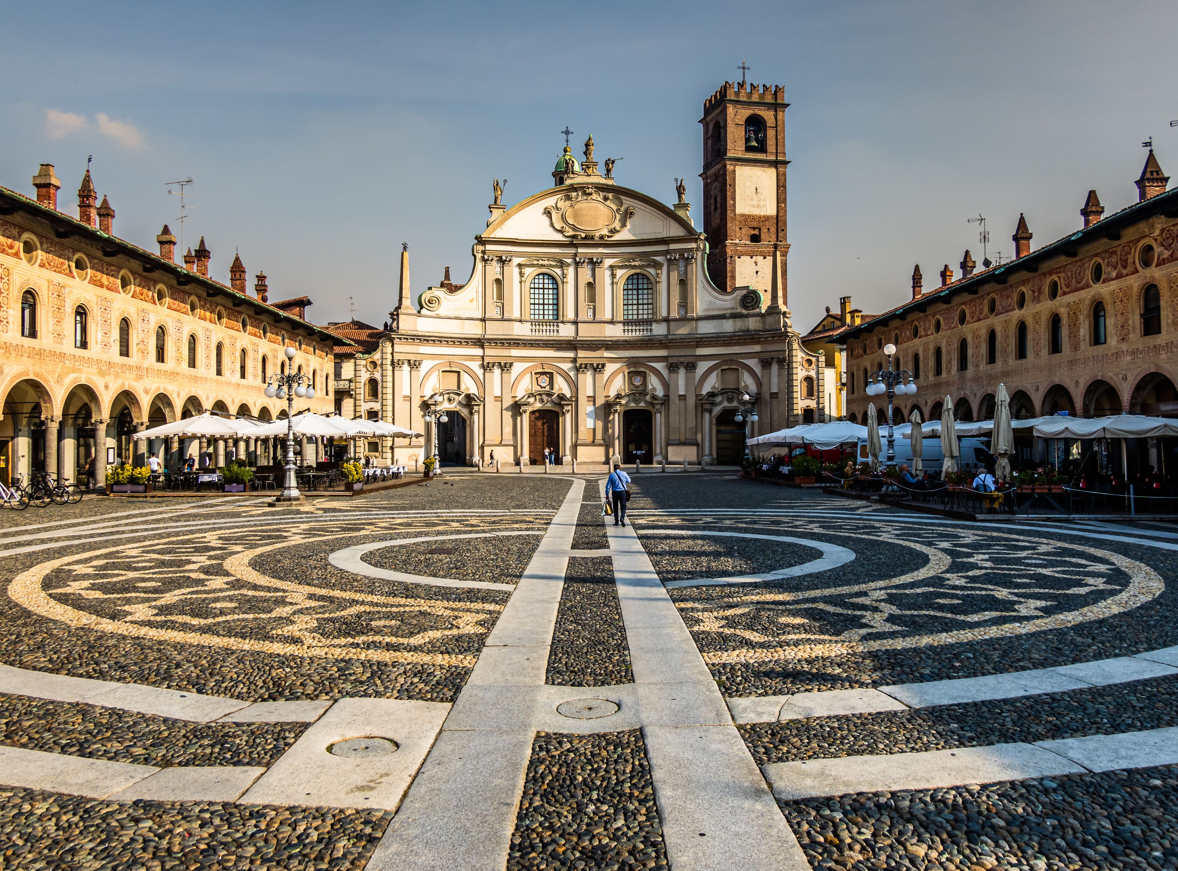 Uma praça histórica com um padrão circular central, uma igreja de destaque com um campanário e edifícios ao redor com mesas ao ar livre.
