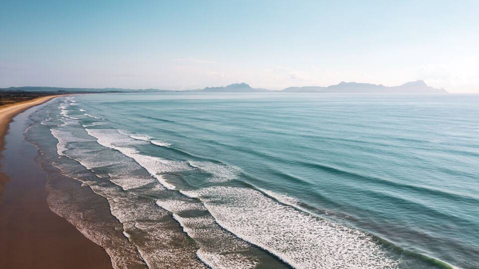 A beach with waves lapping onto the shore and distant mountains under a clear sky.