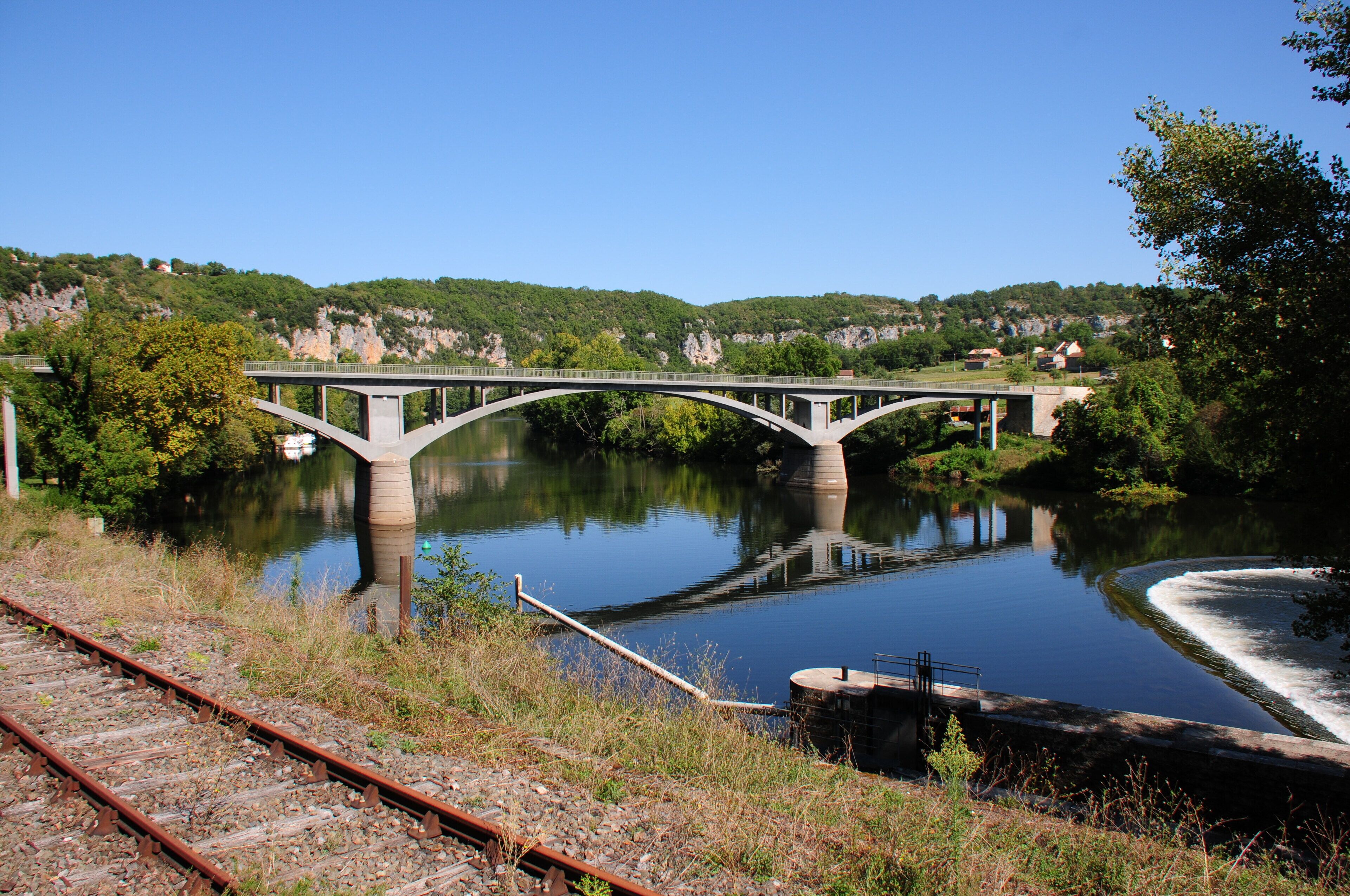 A bridge over a calm river with surrounding greenery and hills.