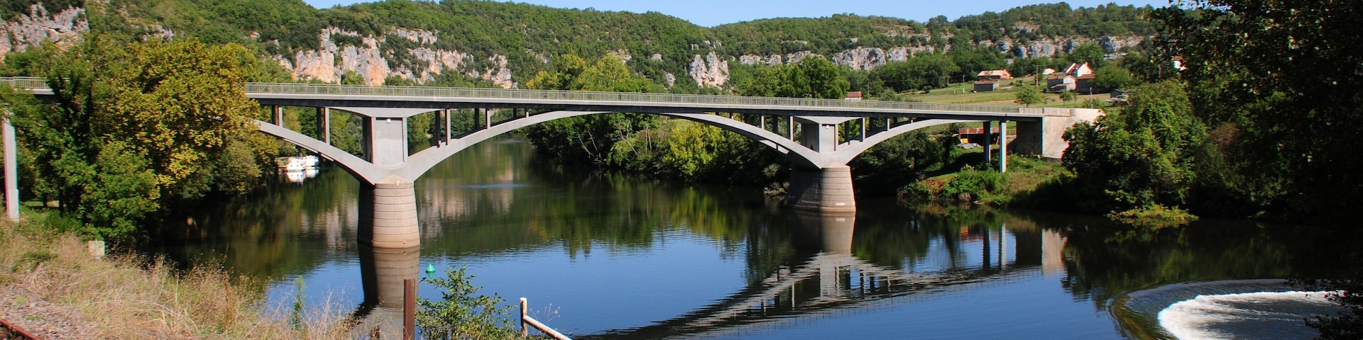 A bridge over a calm river with surrounding greenery and hills.