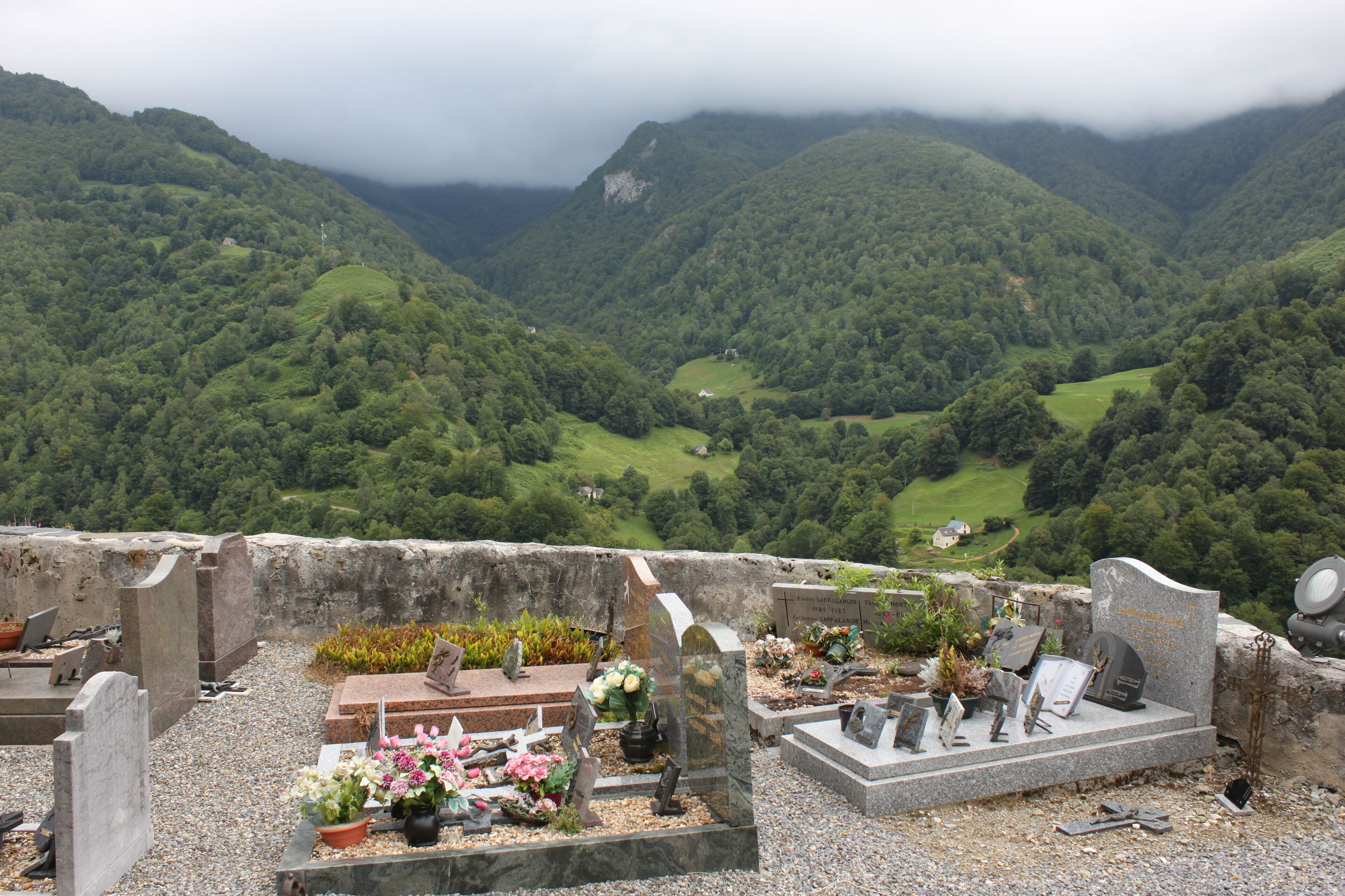 A cemetery with gravestones and flowers, surrounded by lush green hills and a cloudy sky.