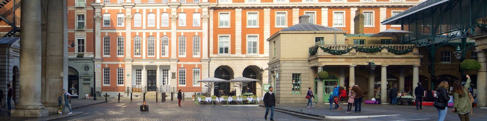 A cobblestone square with historic buildings, a covered walkway, and people walking.