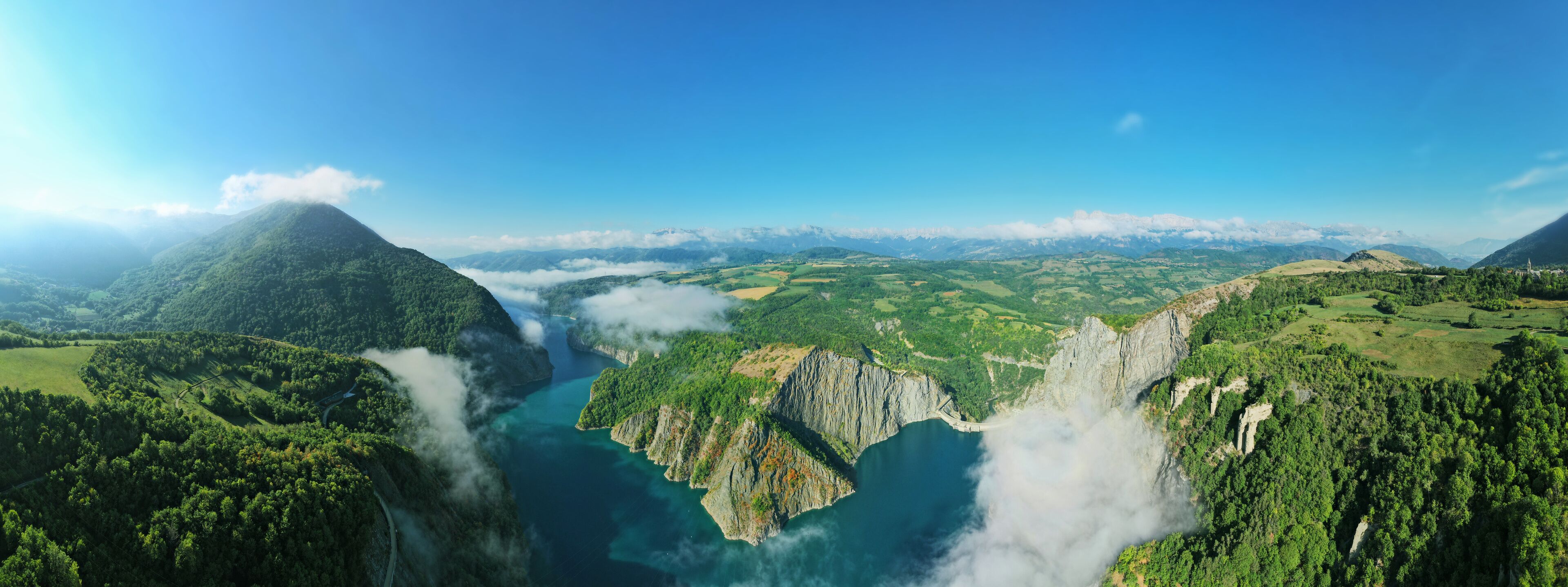 A mountainous landscape with a river, lush greenery, and distant snow-capped peaks.