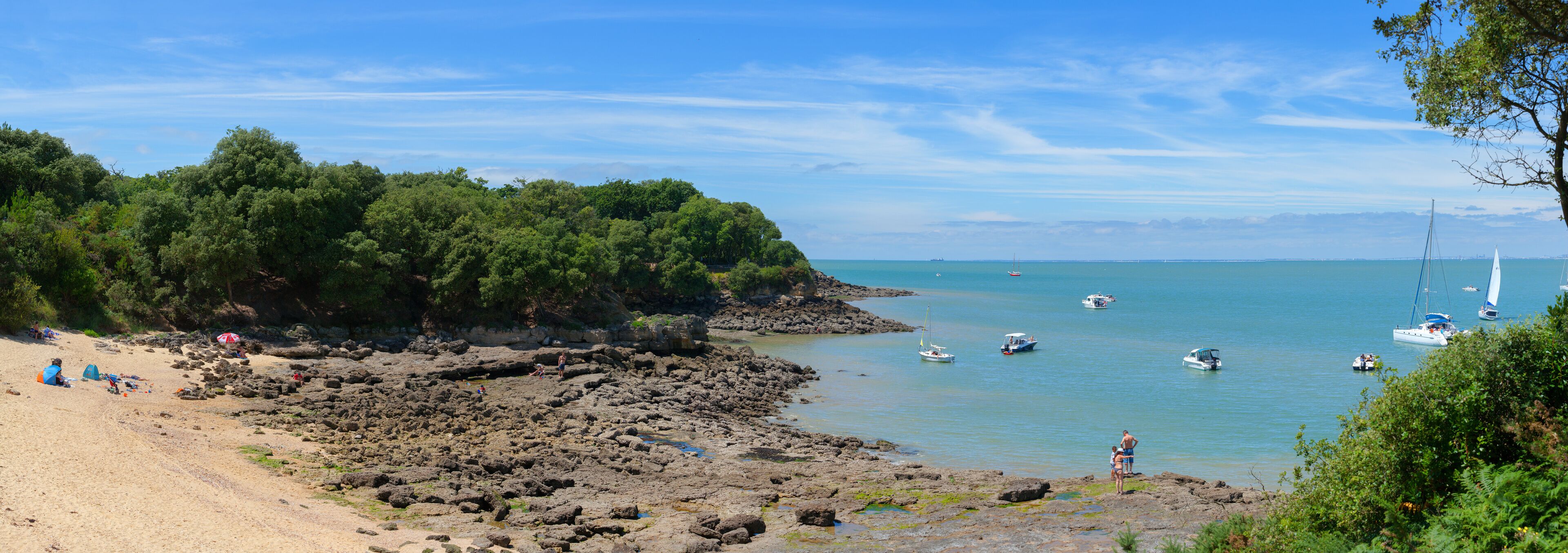 A rocky beach with boats on calm water, surrounded by dense green trees.