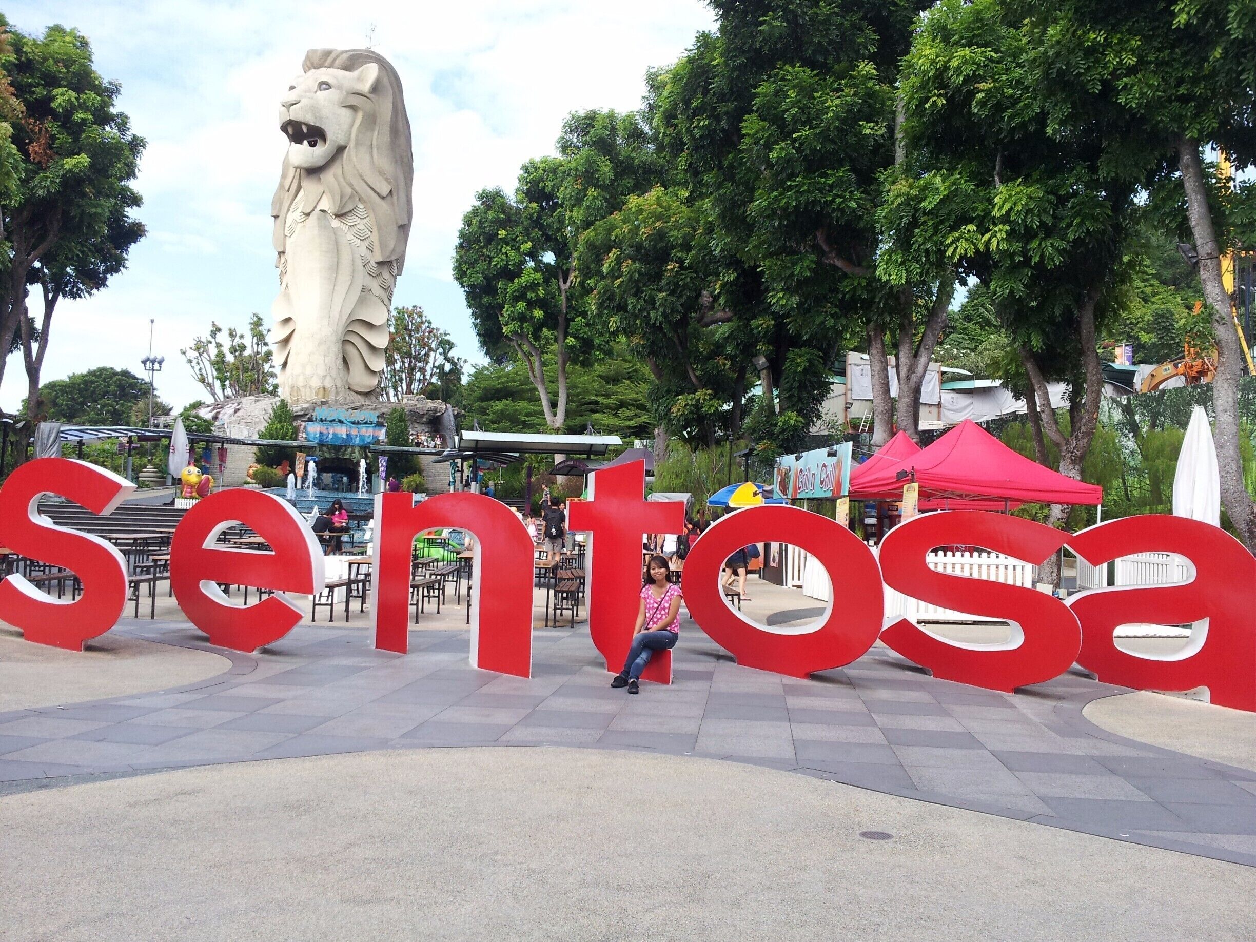 Una estatua grande de un león, señalización roja con la palabra 'Sentosa' y un puesto de comida al fondo.