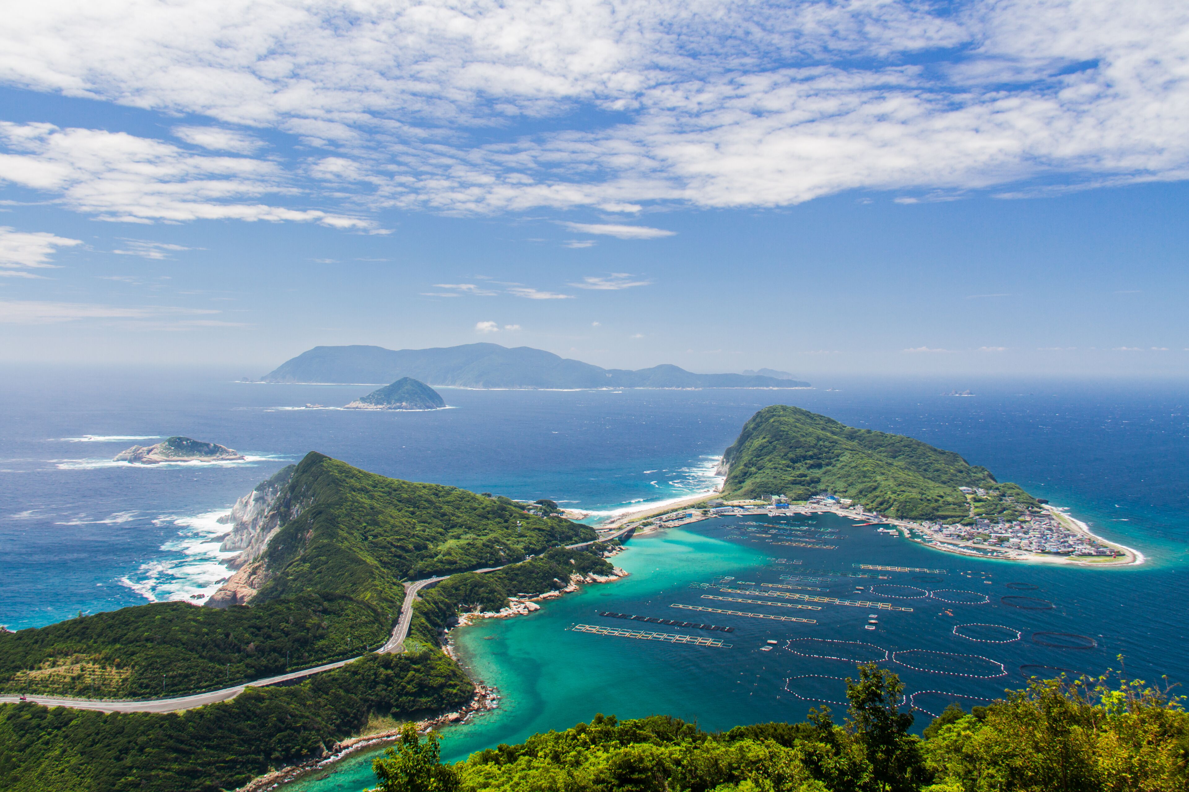 A coastal landscape with lush green hills, a clear blue sea, and a distant mountain range.
