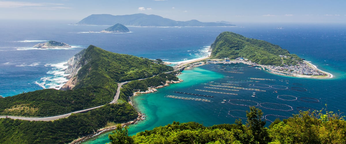 A coastal landscape with lush green hills, a clear blue sea, and a distant mountain range.