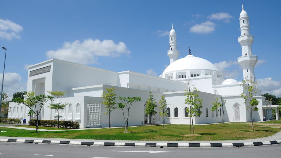 A white mosque with two minarets and a dome, surrounded by a well-maintained lawn and young trees.