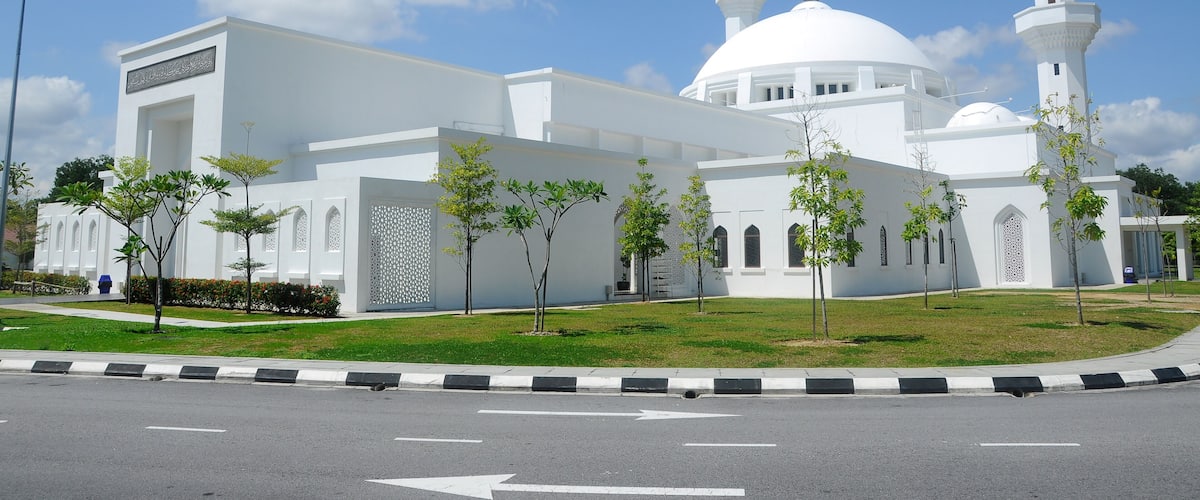A white mosque with two minarets and a dome, surrounded by a well-maintained lawn and young trees.