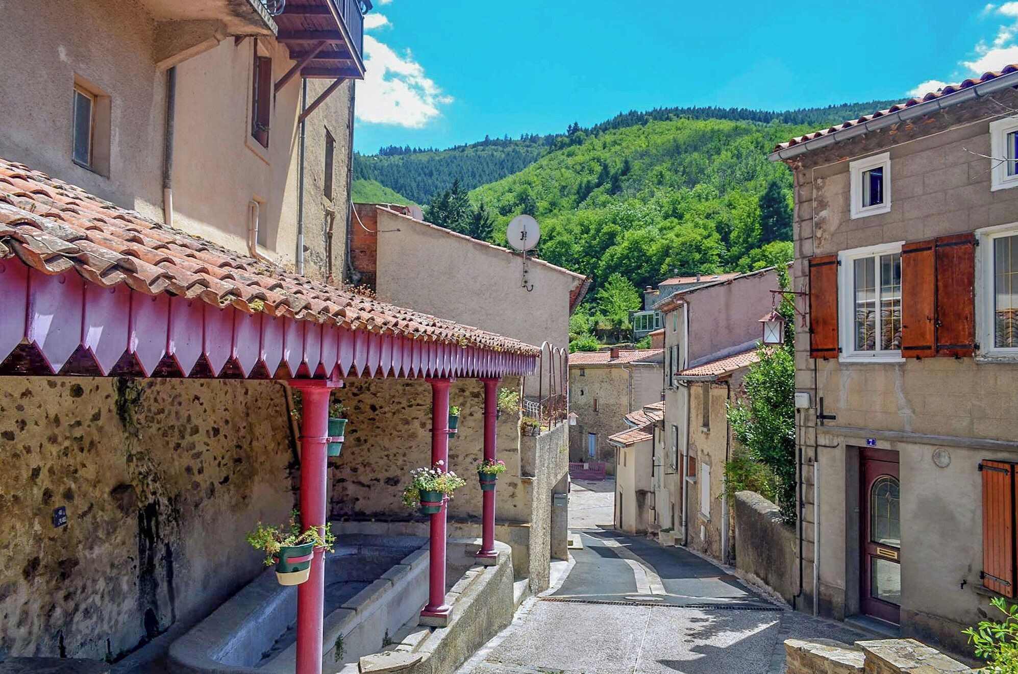 A narrow street with terracotta-tiled roofs, stone buildings, and a lush green hillside in the background.