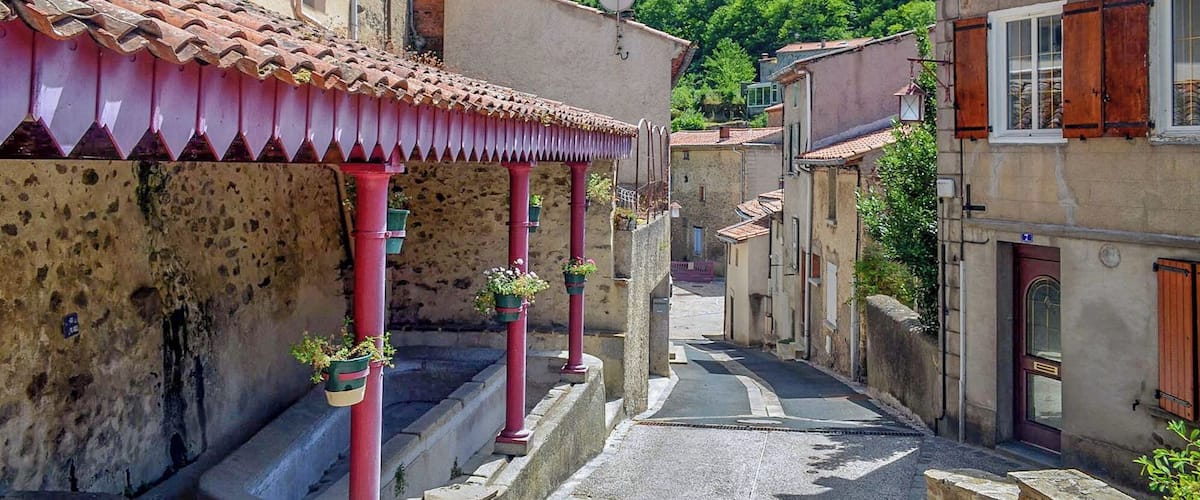 A narrow street with terracotta-tiled roofs, stone buildings, and a lush green hillside in the background.