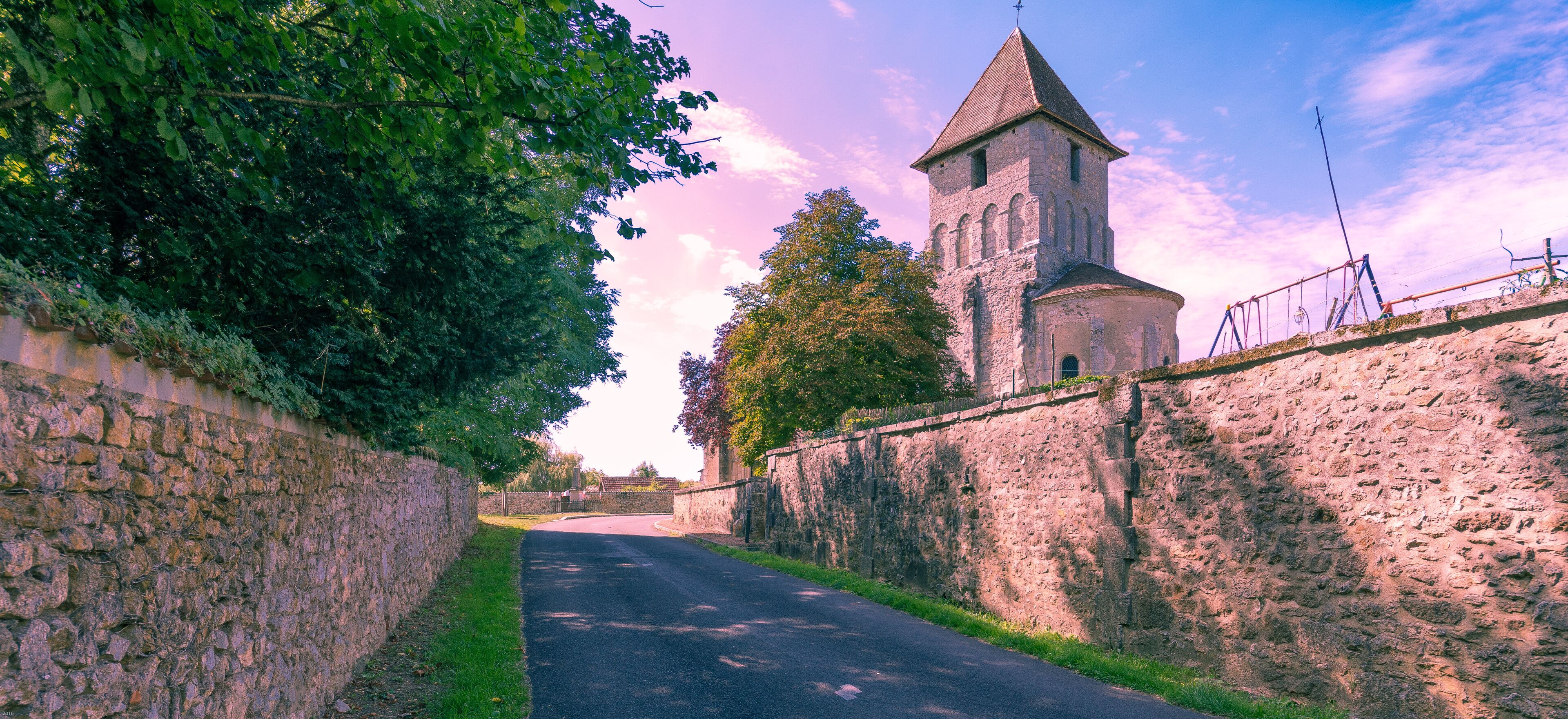 A narrow road flanked by stone walls, leading to a historic tower with a conical roof.