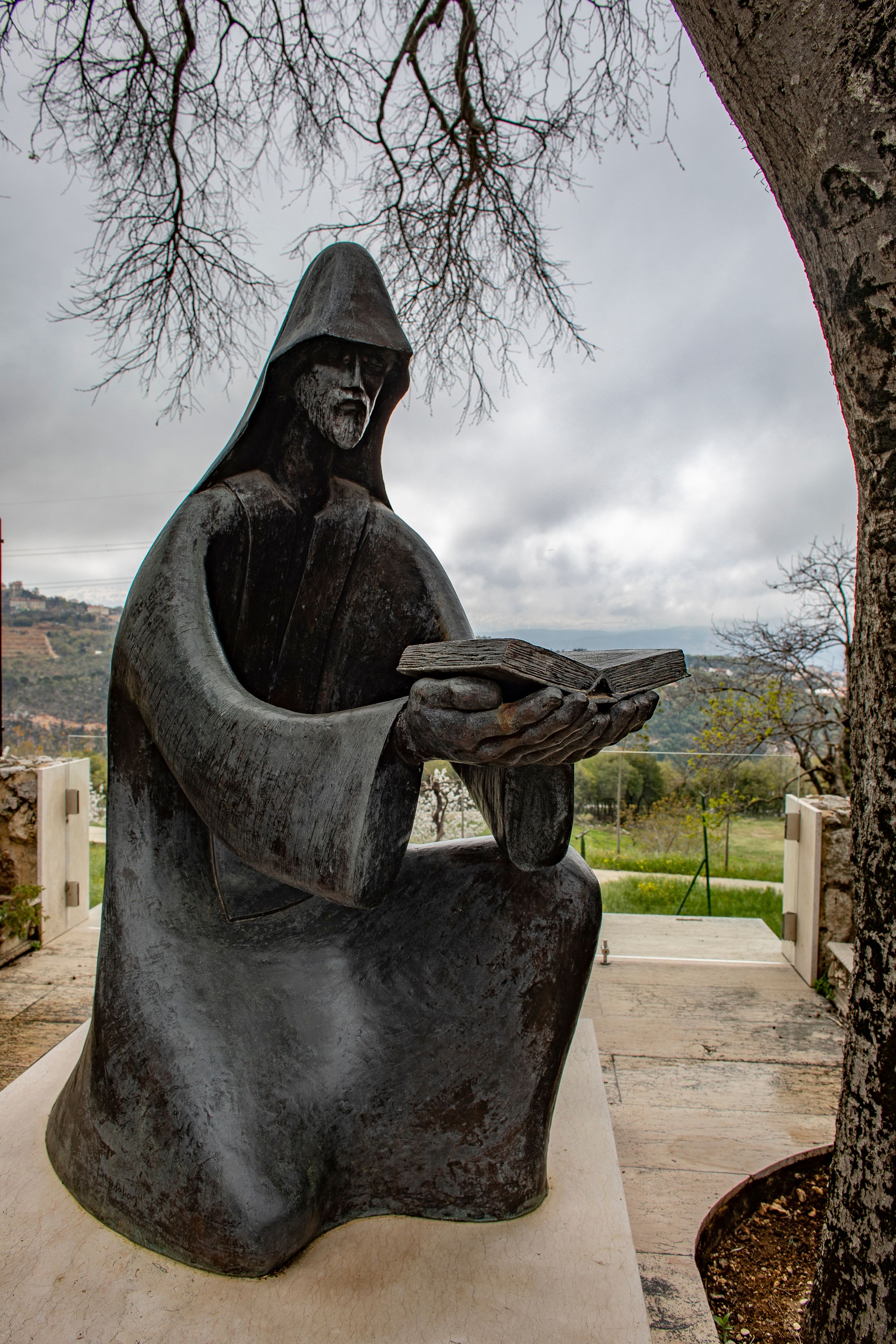 A statue of a figure in a hooded cloak holding a book, seated on a bench with a tree and a view of hills in the background.