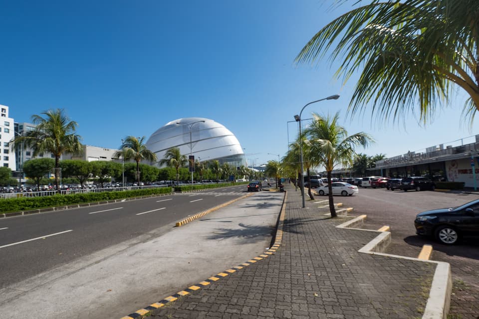 A wide urban street with palm trees, a modern spherical building, and parked cars.