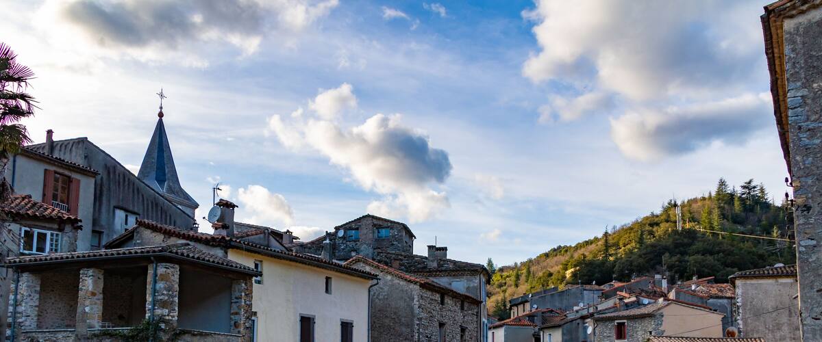 A historic village with stone buildings, a narrow waterway, and a hill with trees in the background.