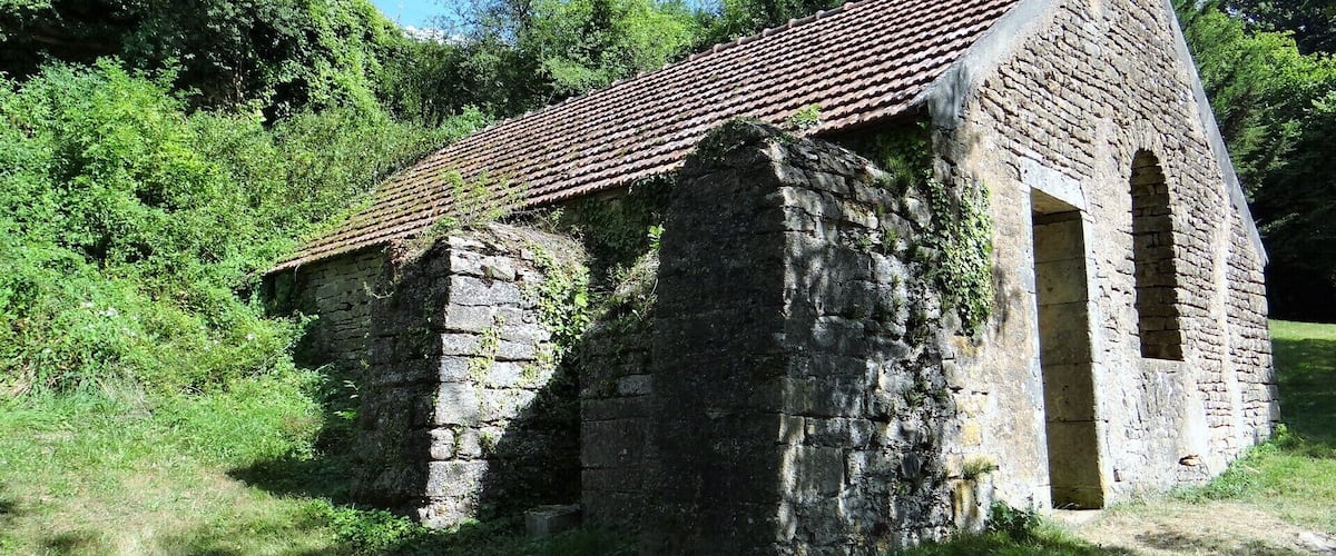 An old stone building with a tiled roof and arched doorway, surrounded by lush greenery.
