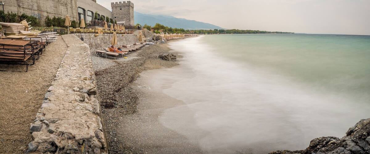 A coastal promenade with a stone wall, beach umbrellas, and a castle-like tower.
