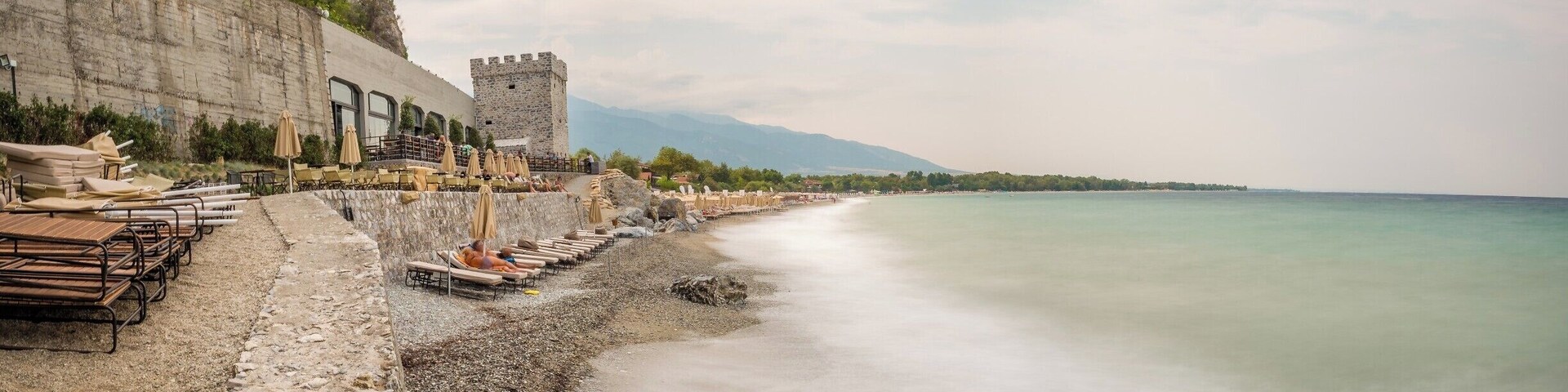 A coastal promenade with a stone wall, beach umbrellas, and a castle-like tower.