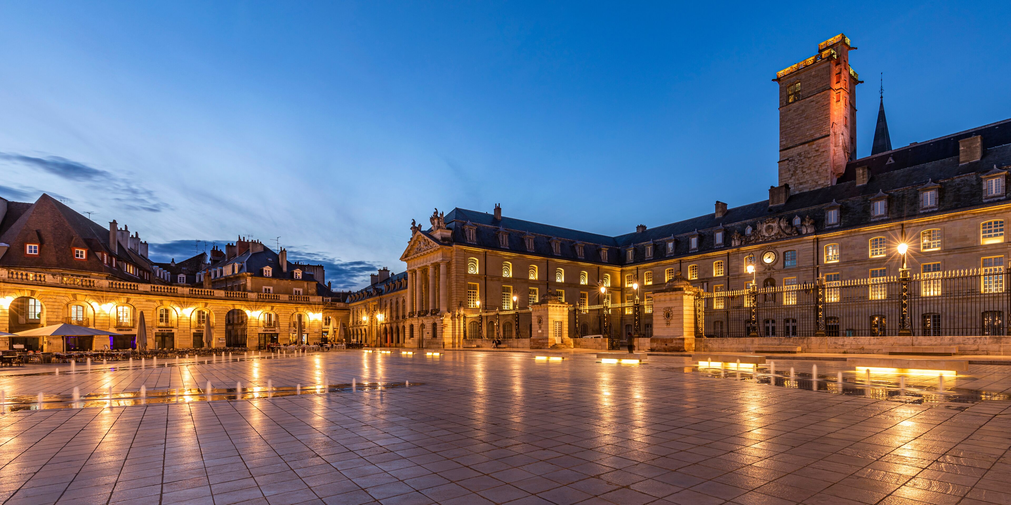 A historic square with a fountain, illuminated buildings, and a clock tower.