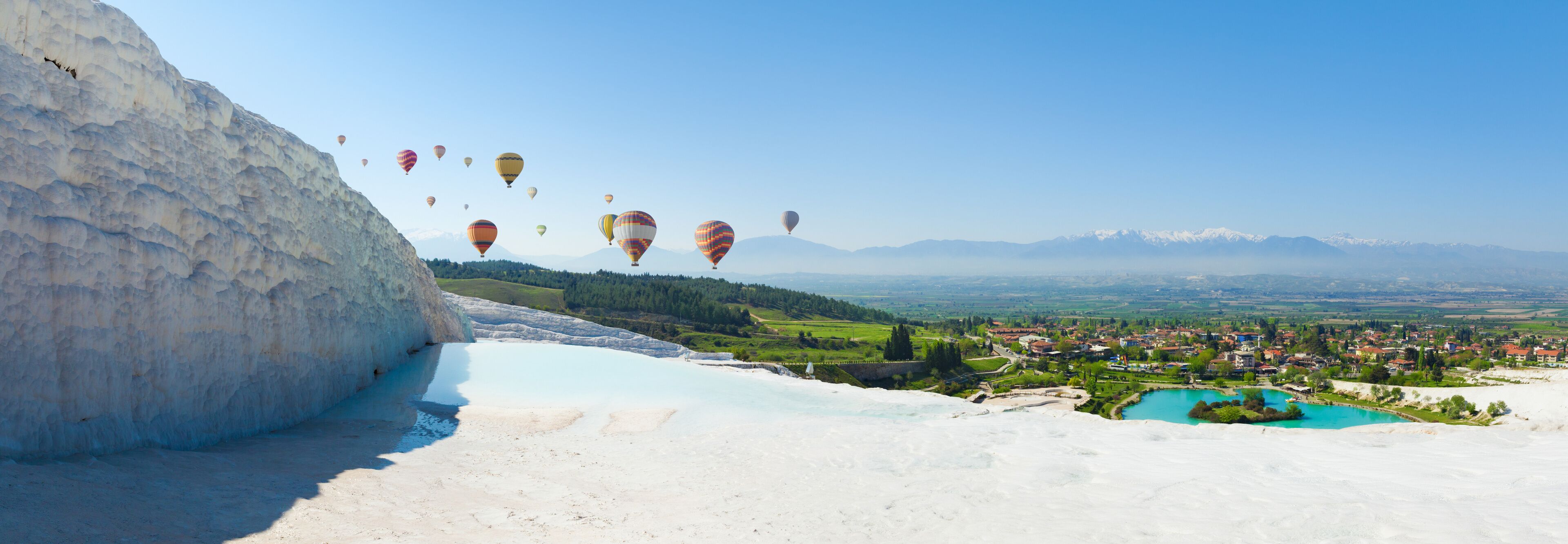A valley with hot air balloons, a town, and mountains in the background.