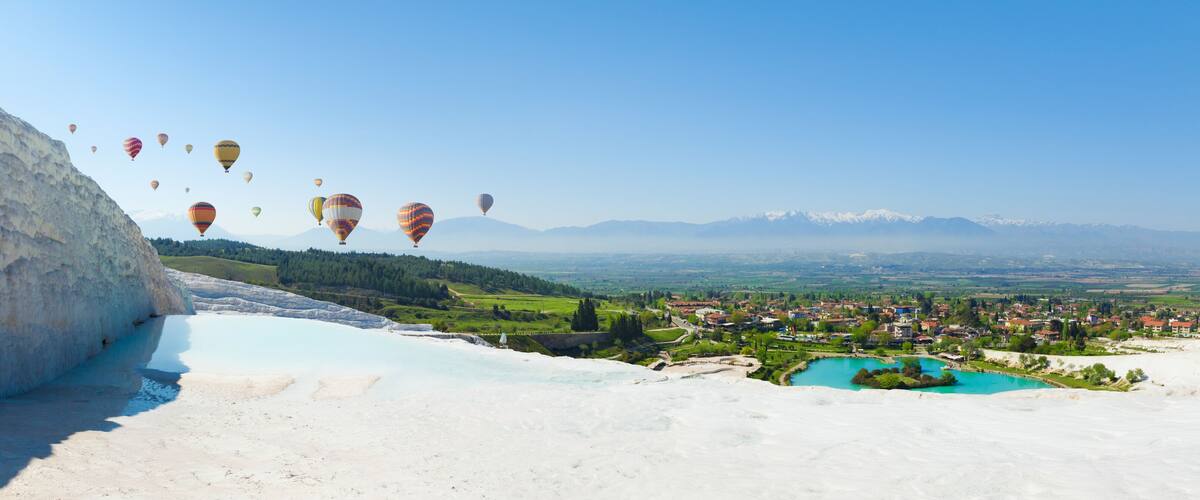 A valley with hot air balloons, a town, and mountains in the background.